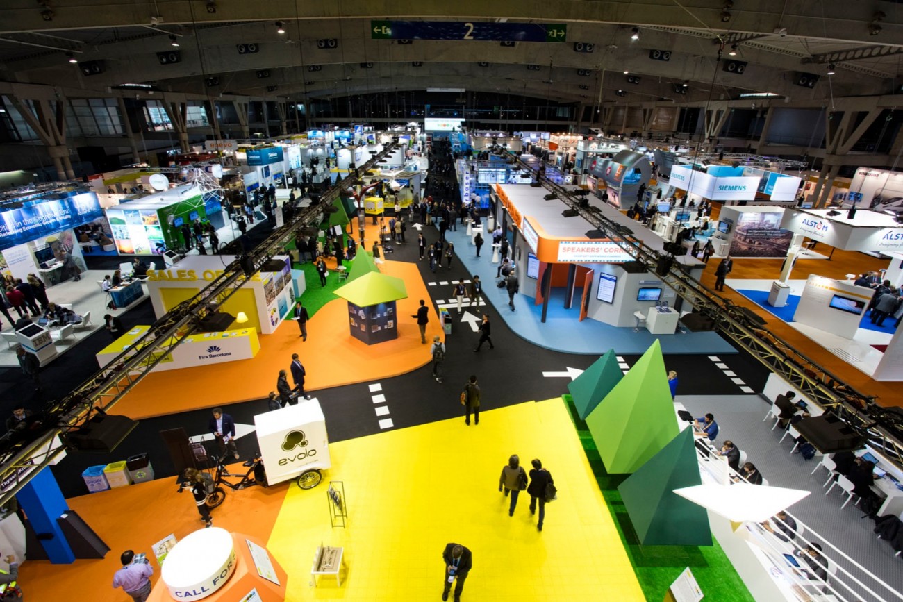 A wide, overhead view of a Smart City exhibition hall, with brightly colored booth areas in yellow, orange, green, and blue, and many attendees walking between technology company stands.